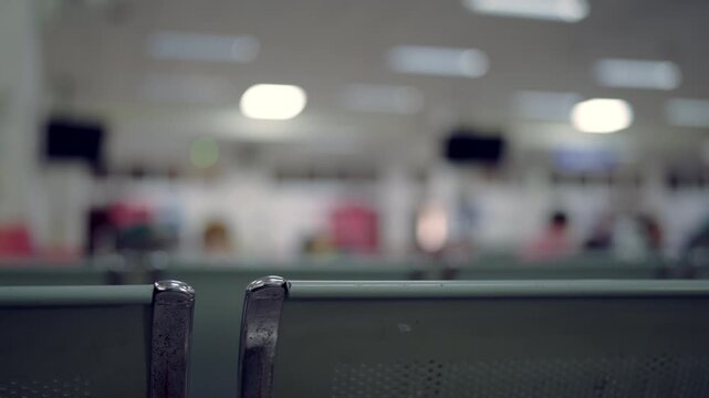 Blurry background shows waiting area. Metal bench frame in foreground. Soft light illuminates distant figures. Indoor space feels quiet and empty. People are seated but blurred
