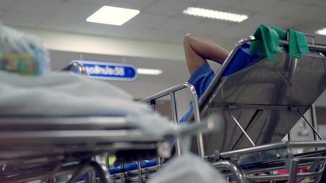 Hospital scene with a person resting on a stretcher. Metal frame and blue padding visible. Background includes signage and overhead lights. Setting implies medical or emergency environment