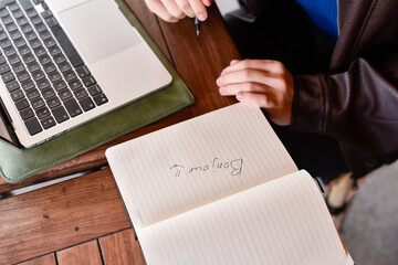 High angle view of a person sitting at a wooden table with a laptop and a notebook. The word Bonjour is written in the notebook next to a pen. © dsbedarieva