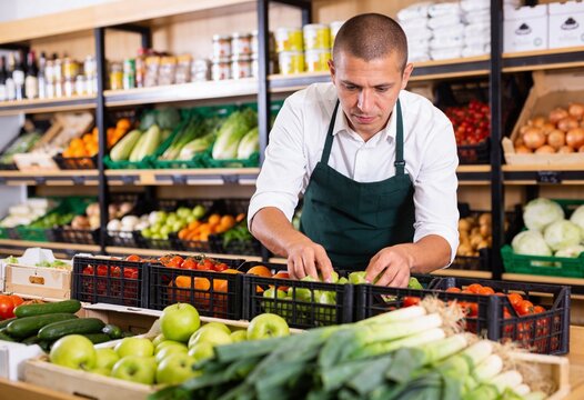 Male grocery store worker in green apron arranging fresh vegetables and fruit in supermarket, retail staff stocking produce for healthy food shopping and small business service