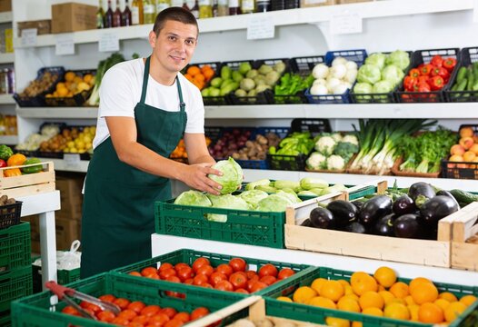 Smiling male grocery store worker in green apron holding a fresh cabbage, supermarket employee arranging organic vegetables and fruits in local market produce section