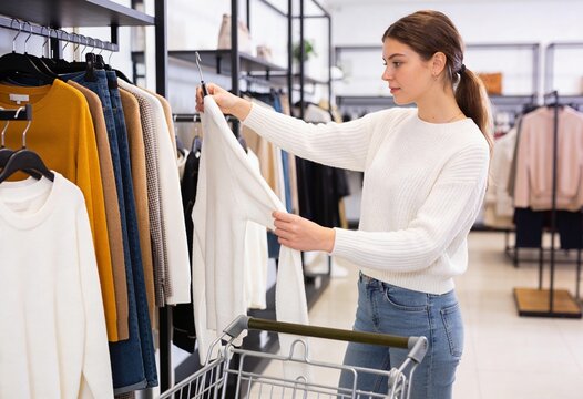 Young woman shopping for clothes in a retail store, holding a white sweater and looking at the fabric, consumerism and fashion lifestyle for retail business and apparel sales