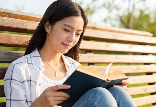 Young Asian woman reading a book while sitting on a wooden bench in a park during golden hour, peaceful outdoor leisure and education for student lifestyle or mental wellness