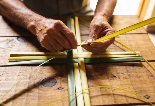 Senior hands weaving a traditional palm leaf cross on a rustic wooden table, handmade religious craft for Palm Sunday celebration and Easter spiritual ritual
