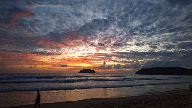 A breathtaking view of the sun setting over the ocean, casting warm golden hues across the waves and clouds while people enjoy the tranquil shoreline. Poo island Kata beach Phuket
