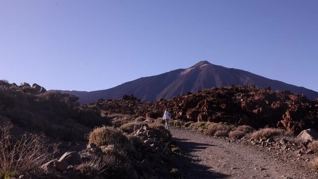 Wide cinematic shot of lone woman in white standing small against massive Mount Teide volcano and dramatic red lava field in Tenerife under deep blue sky. Epic scale travel footage.