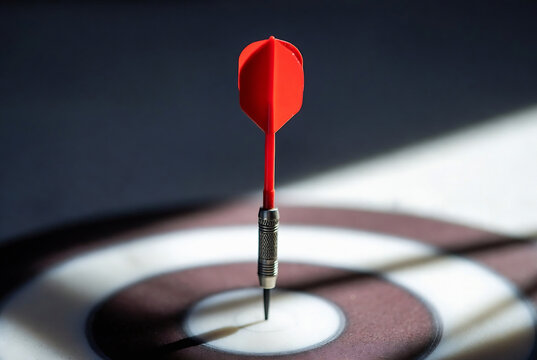 A red dart is aimed at a dartboard target. It&rsquo;s set on a dark surface with light highlighting the target and dart.