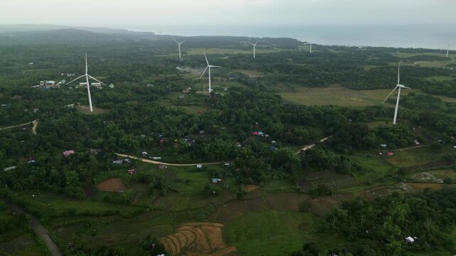 Rising aerial view of wind turbines across lush green fields with distant coastal landscape in Guimaras, Philippines during daytime.