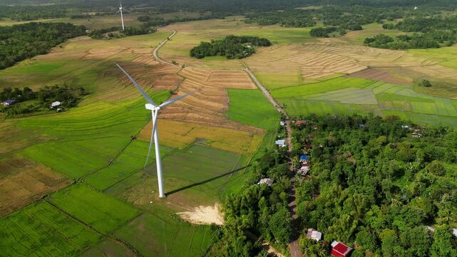 Aerial flyover of a wind turbine standing over patterned farmland and a forested rural village in Guimaras, Philippines during daytime.