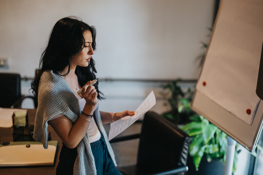 A student reads notes while preparing to present at a flip chart. She reviews her script and rehearses in a quiet classroom setting.