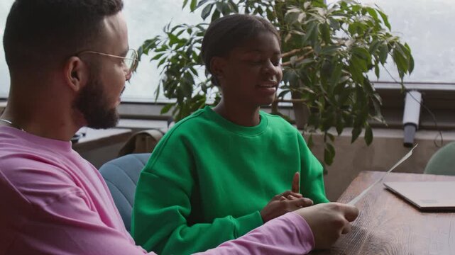 Smiling African American girl and multiethnic young man deciding what to eat for lunch in restaurant while sitting at table near window
