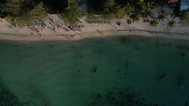 Aerial top-down tracking shot along a quieter section of Koh Lipe beach with longtail boats anchored offshore, beachfront bungalows with colorful roofs, and palm trees lining the white sand shore.