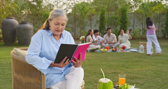 Indian old aged relax woman mom sitting on chair hold book read story novel outdoor house place happy male female man group girl child kid play spend quality time enjoy new day open in air garden lawn