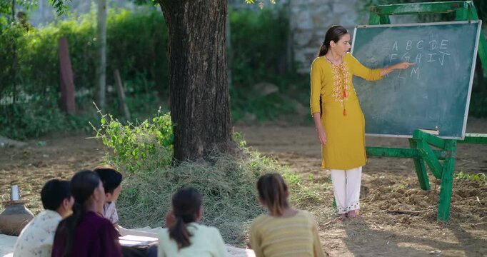 Indian Young adult gen z female tutor standing teach English to villager girl boy outdoor rural area. Happy child group sitting on mat look board learn do study new day time outside ground place