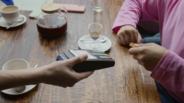 Close-up view of unknown man in pink long sleeve tapping bank card on payment terminal brought to him by anonymous waitress while he sitting at table in cafe