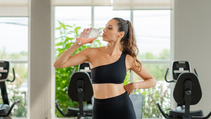 Fototapeta premium Athletic woman refilling water bottle post-exercise. Healthy athletic woman drinking protein shake and using smartphone during exercise break, sports nutrition and active lifestyle.