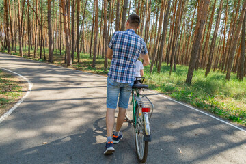Fototapeta premium Dad rides his daughter in a bicycle seat in forest. A man carries a small child on a bicycle.
