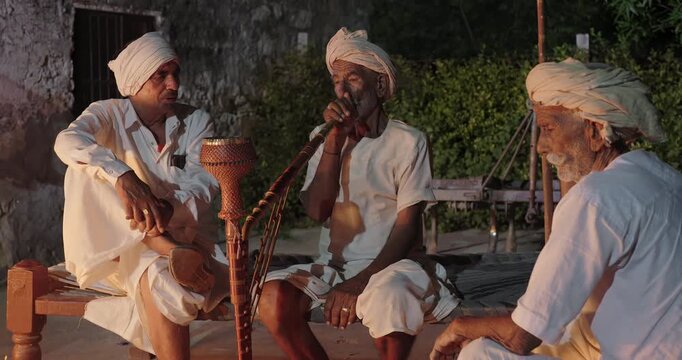 Indian old aged man wear white kurta pajama sitting on woven cot bed enjoy desi life at rural area. Happy four male group talk speak share gossip do hookah hold pipe spend night time village place