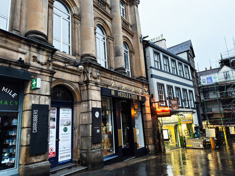 Rainy historic street with shops and stone architecture in Edinburgh, Scotland, UK, featuring Heddle and Reed storefront and wet pavement reflections