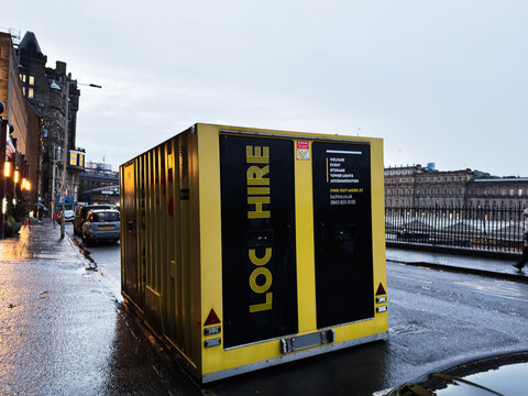 Yellow hire container on a wet city street in Edinburgh, Scotland, UK with waterfront buildings and evening reflections