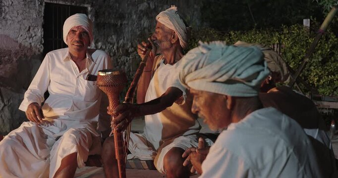Indian old aged man wear white kurta pajama sitting on woven cot bed enjoy desi life at rural area. Happy four male group talk speak share gossip do hookah hold pipe spend night time village place