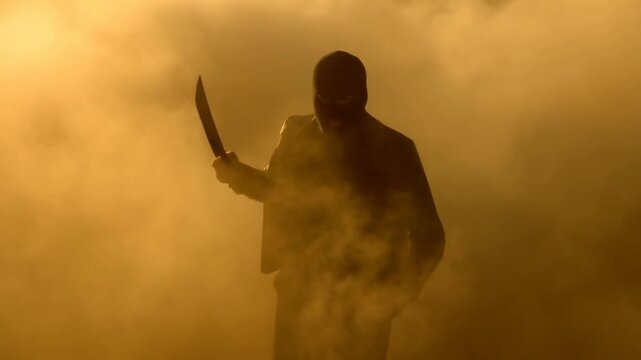 Tracking shot of masked man walking forward with raised machete through dense smoke filled studio haze backlit golden silhouette approaching camera