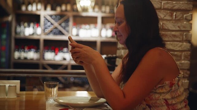 Woman sits alone at table in elegant restaurant or wine bar, using her smartphone. She is waiting for her order, browsing online or texting. Cozy, warm atmosphere with blurred background