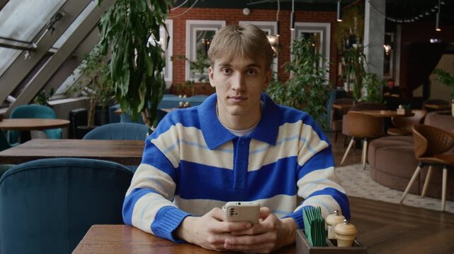 Portrait of Caucasian gen Z man wearing white and blue sweater looking at camera using smartphone at restaurant table during lunch time