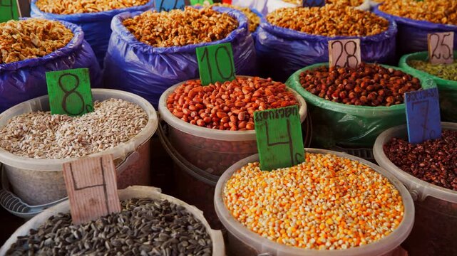 Camera pans across a vibrant market stall. Buckets and bags are filled with a colorful assortment of nuts, seeds, and grains like corn, peanuts, and sunflower seeds. For sale at a local bazaar