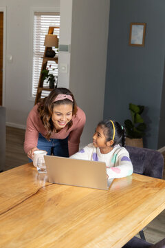 Mother and daughter leaning over dining table, working on laptop with white mug, water glass
