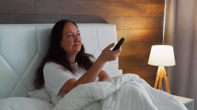 Middle aged woman relaxes in bed, watching television. Her facial expressions change from happy to serious and back to smiling as she uses remote control to switch channels or watch a movie