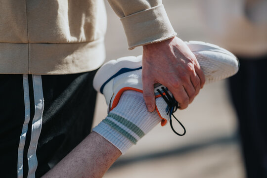 A young athlete stretches a raised leg while holding a sneaker with one hand. The image shows an outdoor warm-up moment before training or a sports activity.