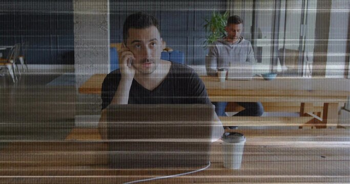 Talking man wearing black T-shirt holding phone at cafe table, with open laptop and coffee cup