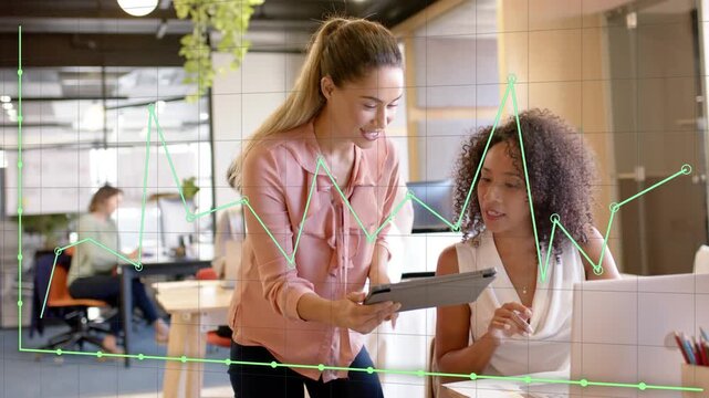 Standing woman showing tablet to seated coworker for business, green graph annotating tablet data