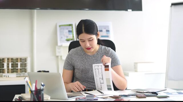 Asian Interior designer sitting in front of laptop talking with client on video call, showing architecture floor plan blueprint of residence and pantone. meeting and online conference.