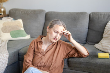 Obraz premium Frustrated woman sitting nearby sofa and feeling stressed after trying to remove a visible stain from a gray sofa using a sponge.. Home cleaning problem and everyday domestic frustration concept.
