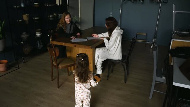 Young daughter in pajamas carrying a toy approaches her working mother, who is busy in a meeting with another woman, representing the challenges of balancing work and family life
