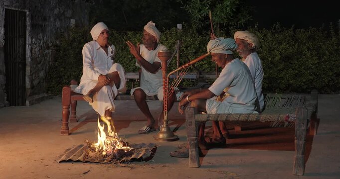 Indian old aged man wear white kurta pajama sitting on woven cot bed enjoy desi life at rural area. Happy four male group talk speak share gossip do hookah hold pipe spend night time village place