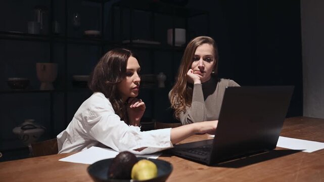 Two businesswomen discussing work on a laptop in a dark office at night, one colleague pointing at the screen and explaining a concept while the other listens attentively to the strategy