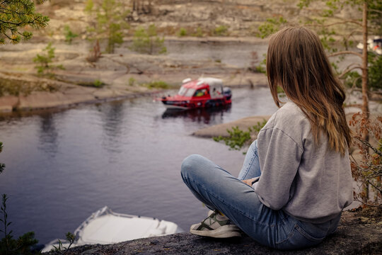 A young woman sits on a high rocky ledge, her back to the camera, overlooking a calm bay where a red motorboat is cruising through a rugged, northern landscape. Sortavala, Karelia, skerries, Ladoga