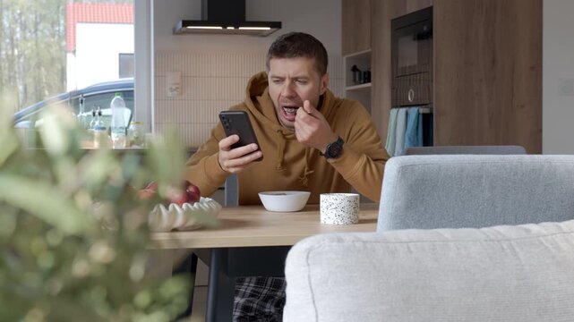 Man eating oatmeal porridge and looking at smartphone while sitting at kitchen table. Person using phone during breakfast.