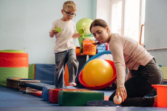 Young boy in glasses plays with colorful blocks while woman assists him in a bright indoor playroom filled with various toys and equipment for children