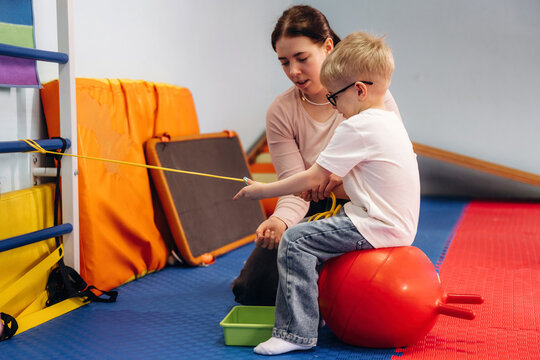 Female therapist assists young boy with special needs using a red therapy ball and green container in a colorful indoor playroom with soft flooring and gym equipment