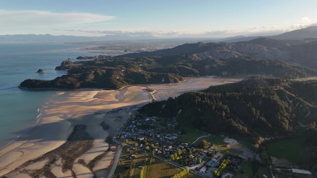Panoramic aerial view of Marahau town and Abel Tasman coastline at sunset, South Island, New Zealand, with golden tidal sandbanks, turquoise ocean, rolling hills and village in warm evening lights.