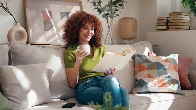 Video of mature beautiful woman reading a book while drinking a cup of coffee sitting on couch at home.