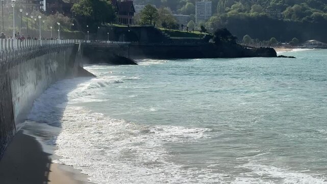 4K sunny footage of waves along Paseo Nuevo seawall, San Sebastian. Historic villa, sandy beach, green hills, tourists on promenade. Warm travel b roll for Basque Country destination content.