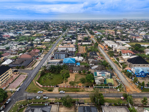 Aerial view of a sprawling urban landscape crisscrossed by grey roads, dotted with diverse buildings and vibrant green spaces under an expansive, cloudy sky, Uyo, Akwa Ibom, Nigeria.