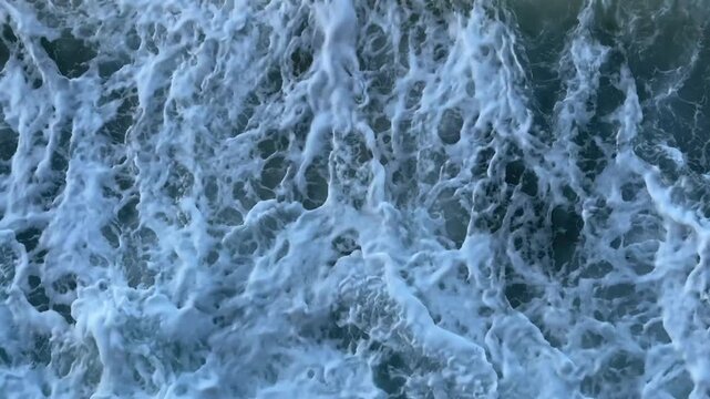 Dramatic 4K aerial overhead video of wave foam trails spreading on dark teal rocky ocean bottom. High-contrast white foam on dark water  ideal for nature, surf, and abstract background content.