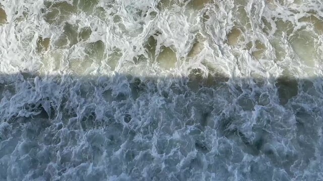 4K aerial overhead video of a wave rolling over sandy shallow ocean bottom. White foam crest divides golden sandy shallows and dark teal water. Ideal for travel, nature, drone footage, and abstract.