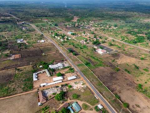 Aerial view of a long, dark road cutting through a patchwork of vibrant green foliage and dry brown earth, dotted with scattered buildings under a bright sky, Elu, Abia, Nigeria.